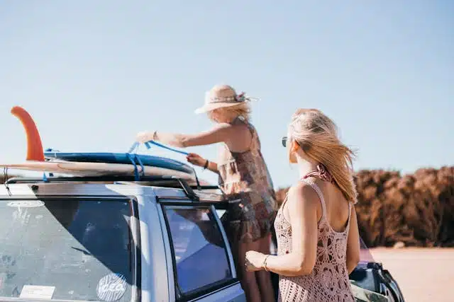 Two women securing a surfboard to the roof of a car on a sunny day (keeping items in the car if possible, rather than on the roof can reduce wind drag and help affordable driving)