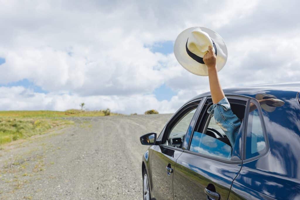 An arm extends from a car window, waving a white hat joyfully against a cloudy sky. Perfect for capturing the spirit of an adventurous girls' weekend away in Australia, exploring gravel roads without breaking the budget.