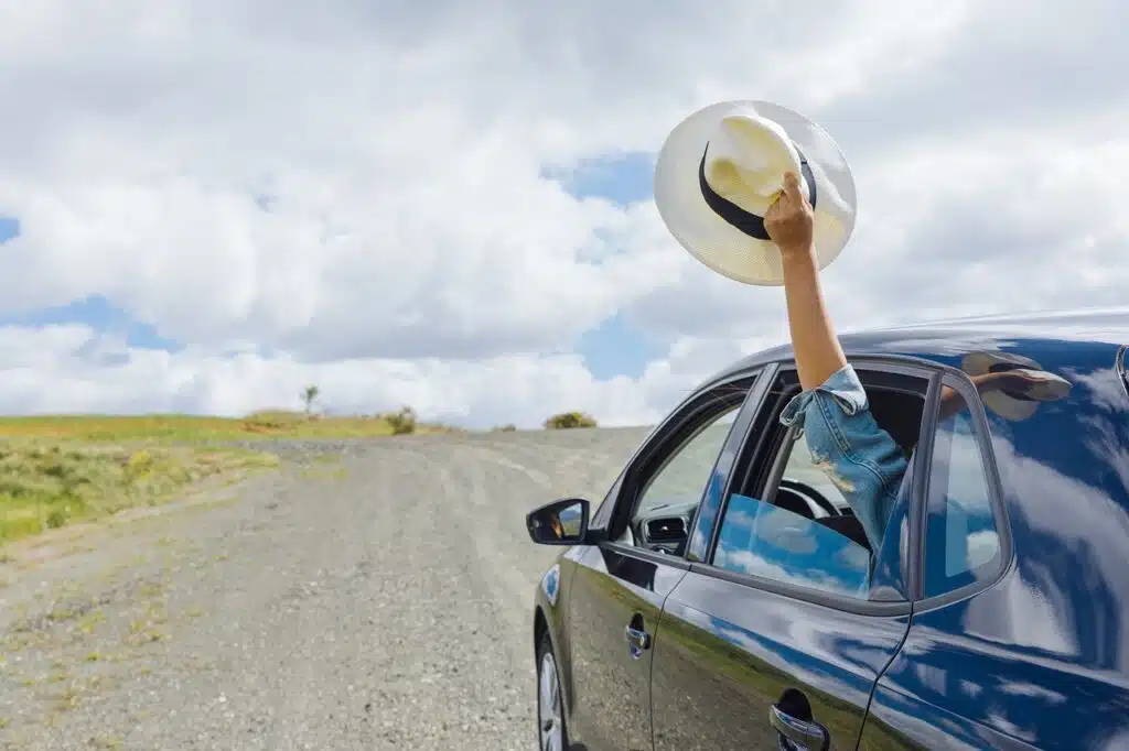 An arm extends from a car window, waving a white hat joyfully against a cloudy sky. Perfect for capturing the spirit of an adventurous girls' weekend away in Australia, exploring gravel roads without breaking the budget.