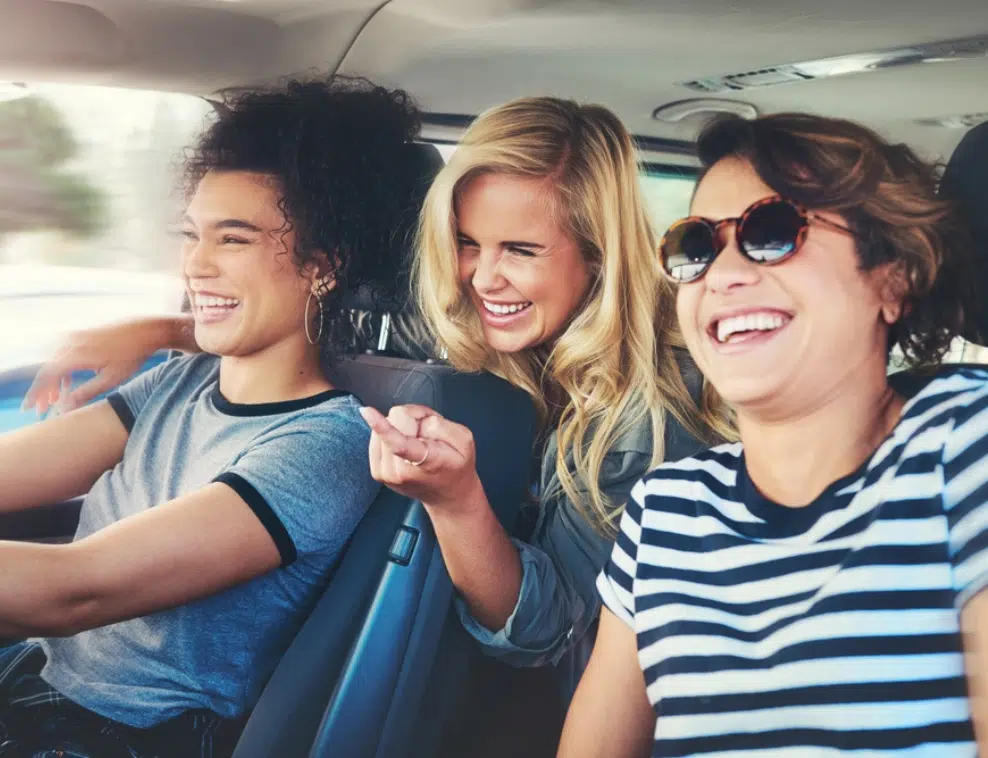 Three friends sitting in a car, smiling and laughing during their girls' weekend away. The driver wears a gray shirt, while the passengers don striped and dark tops. Bright sunlight filters through the windows as they embrace this budget-friendly adventure across Australia.