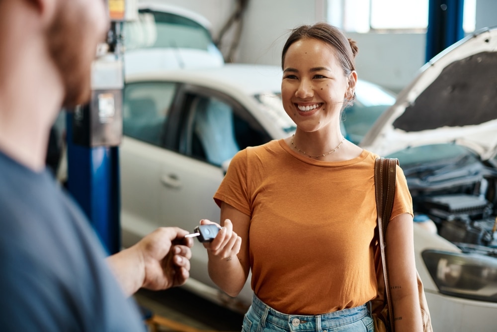 In a garage, a person receives car keys from another, with a white car in the background and its hood open. They're likely discussing how much it costs to service a car or how often you should service your vehicle to keep it running smoothly.