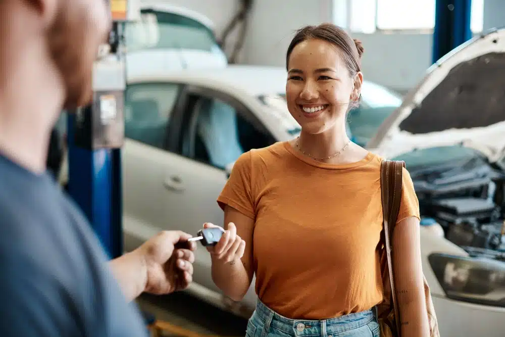 In a garage, a person receives car keys from another, with a white car in the background and its hood open. They're likely discussing how much it costs to service a car or how often you should service your vehicle to keep it running smoothly.