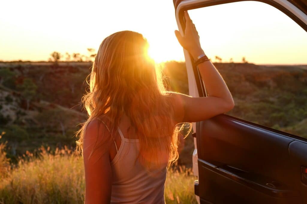 A person with long hair stands by an open car door, gazing at the sunset over a landscape of grasses and distant trees. It's the perfect moment to inspire girls' weekend away ideas in Australia for those seeking adventure and tranquility on a budget.