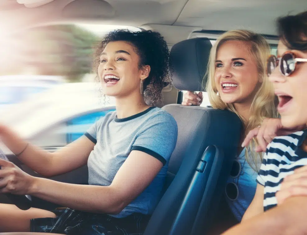 Three women in casual attire are smiling and laughing inside a moving car. The driver is focused on the road while the two passengers, brimming with excitement for their girls' weekend away, cheerfully look out. Sunlight filters through the window as they explore Australia on a budget.