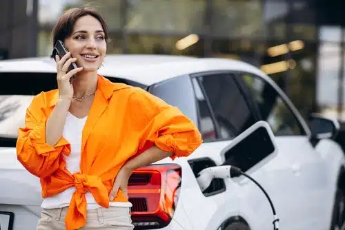 A woman in an orange shirt asks electric car questions on a phone while standing by a white electric car that is charging.