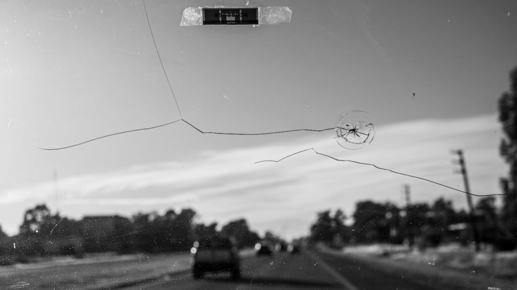 Cracked windshield with visible chip, black and white photo of a road with blurred vehicles and trees. Can you drive a car with a cracked windscreen? The image captures the risk and peril of navigating streets when your view is compromised by windscreen damage.