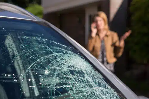 A close-up of a shattered car windshield highlights the danger of windscreen damage. In the background, a person gestures while talking on the phone, perhaps pondering, can you drive a car with a cracked windscreen?.