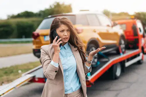 A woman makes a phone call to her mechanic while watching her vehicle get lifted on to a tow truck after a run in with another driver