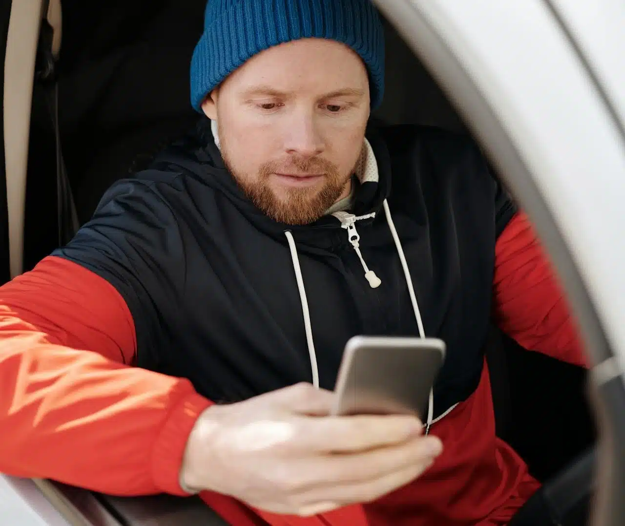 A man in a beanie is using a cell phone to compare car insurance quotes in Australia while sitting in his parked car.