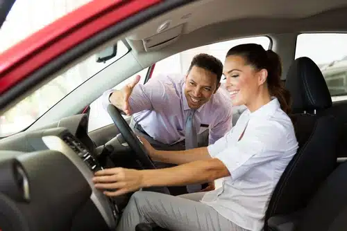 A brunette woman sits in a red car to buy while the salesman shows her features