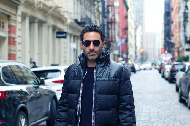 A man wearing a black jacket and sunglasses stands on a cobblestone city street lined with parked cars and buildings, reminding us to reduce car costs and save petrol by choosing to walk more often.