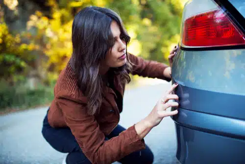 A woman examines the rear bumper of a parked car on a roadway with trees in the background, seemingly contemplating if touch up paint for cars will suffice for car scratch repair.