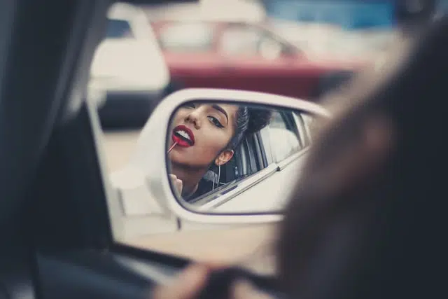 woman doing lipstick makeup in side mirror while driving