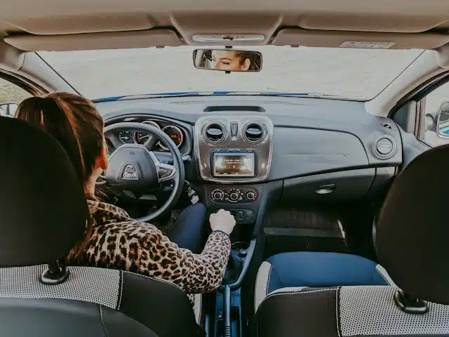 Woman driving a car, viewed from the backseat, adjusting the gear shift with dashboard and steering wheel visible, highlighting the car's safety features.