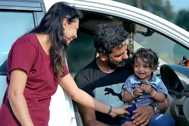A man and a woman smiling at a toddler holding a smartphone, all sitting by an open car door showcasing the safety features of the car in a sunny outdoor setting.