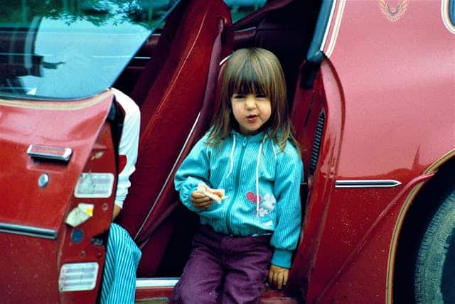 Young girl eating a snack while sitting on the doorstep of a red vintage car, equipped with modern safety features.
