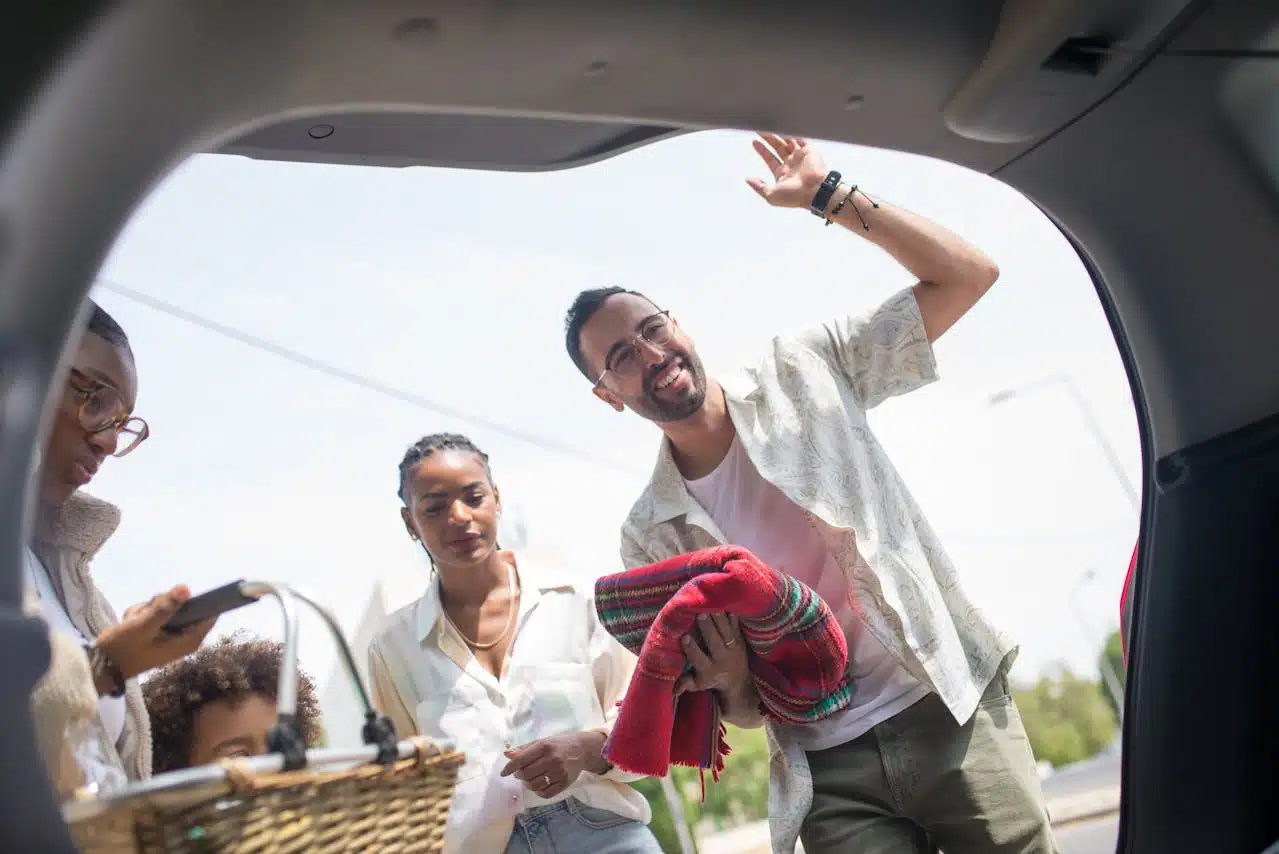 Man loading picnic supplies into a car trunk equipped with safety features, greeting family on a sunny day.