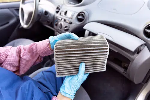 A person wearing blue gloves holds a dirty cabin air filter inside a vehicle.
