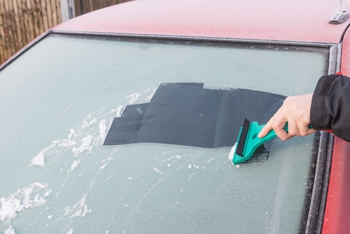 Close up of hand using scraper to take ice off car windscreen. Using a windscreen cover can protect your car from winter ice and snow