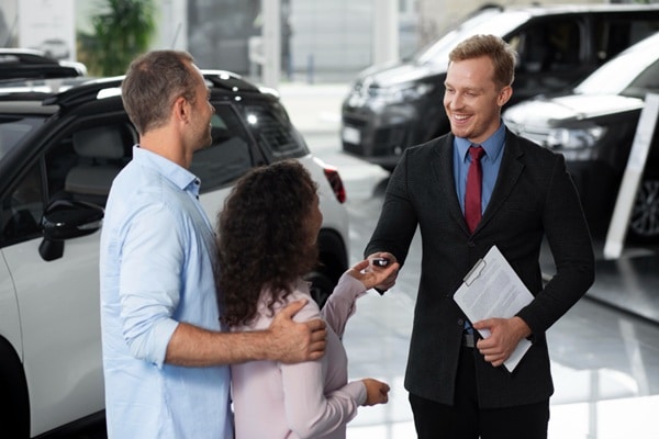 A car salesperson hands keys to a couple inside a car dealership showroom, where cars are on display—highlighting one way an auto broker or car broker can help buyers find the perfect vehicle.