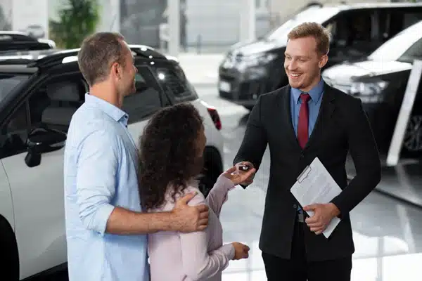 A car salesperson hands keys to a couple inside a car dealership showroom, where cars are on display—highlighting one way an auto broker or car broker can help buyers find the perfect vehicle.