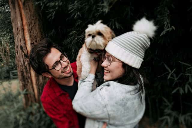 A man and a woman, both wearing glasses, smiling while the woman holds up a small dog. They are outdoors near some trees, celebrating International Dog Day.