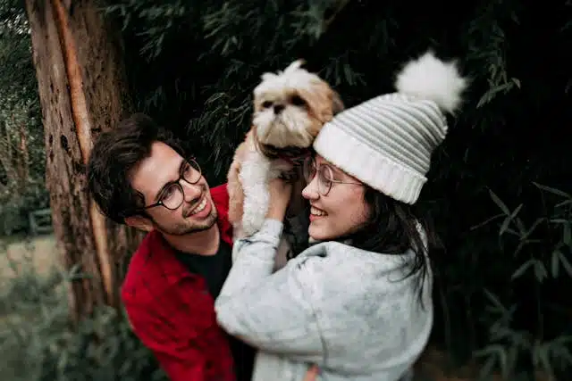 A man and a woman, both wearing glasses, smiling while the woman holds up a small dog. They are outdoors near some trees, celebrating International Dog Day.
