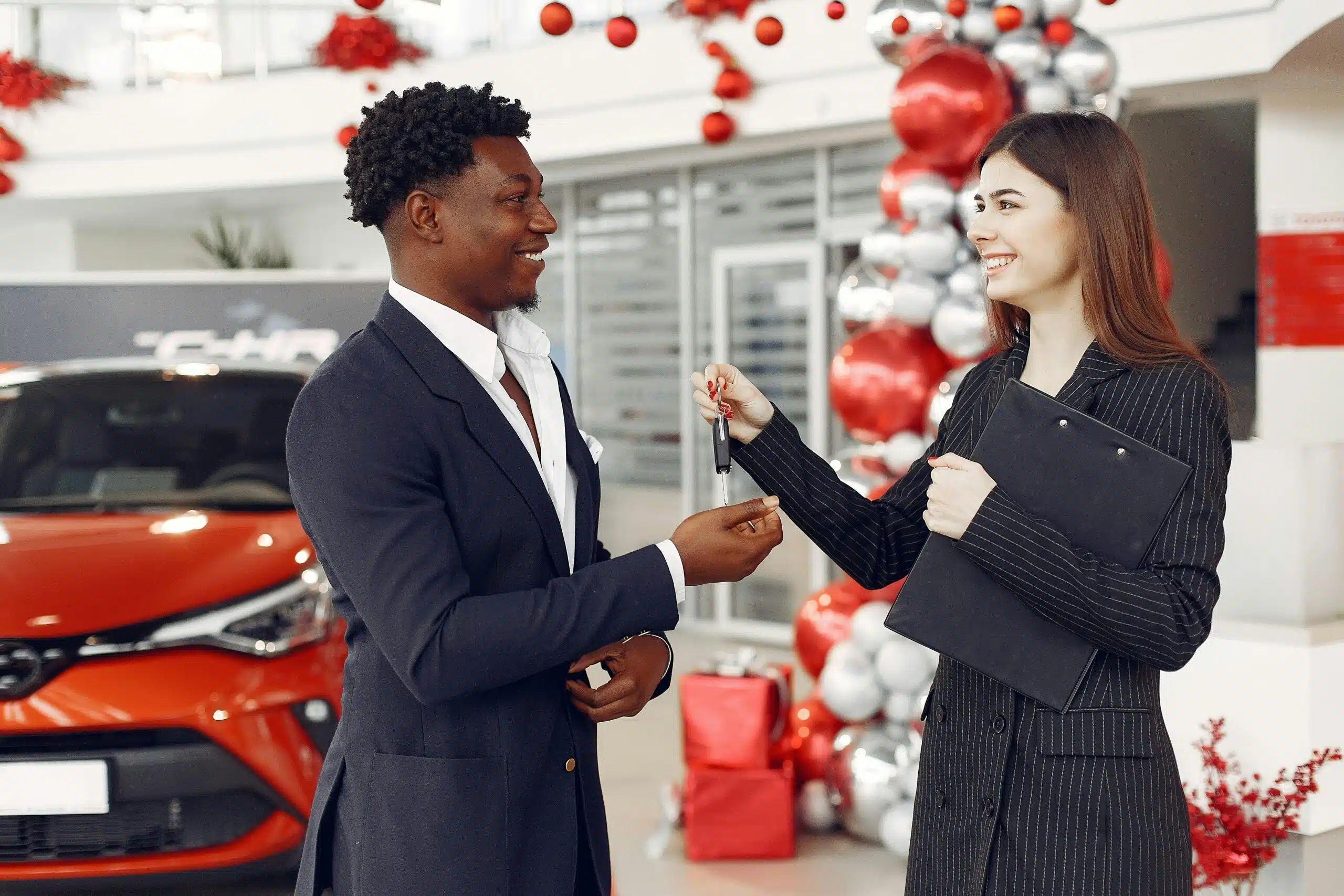 In a festive, ornamented showroom, a person hands over car keys to another, with a shiny red car gleaming in the background. It's the best time to embrace the Christmas spirit and drive away in your new car.