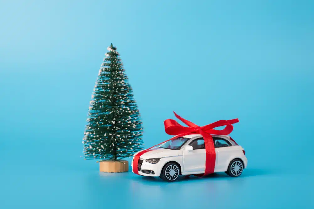 A small toy car with a red bow gleams next to a miniature Christmas tree against a blue background, sparking the thought: is Christmas a good time to buy that new car you've been dreaming about?
