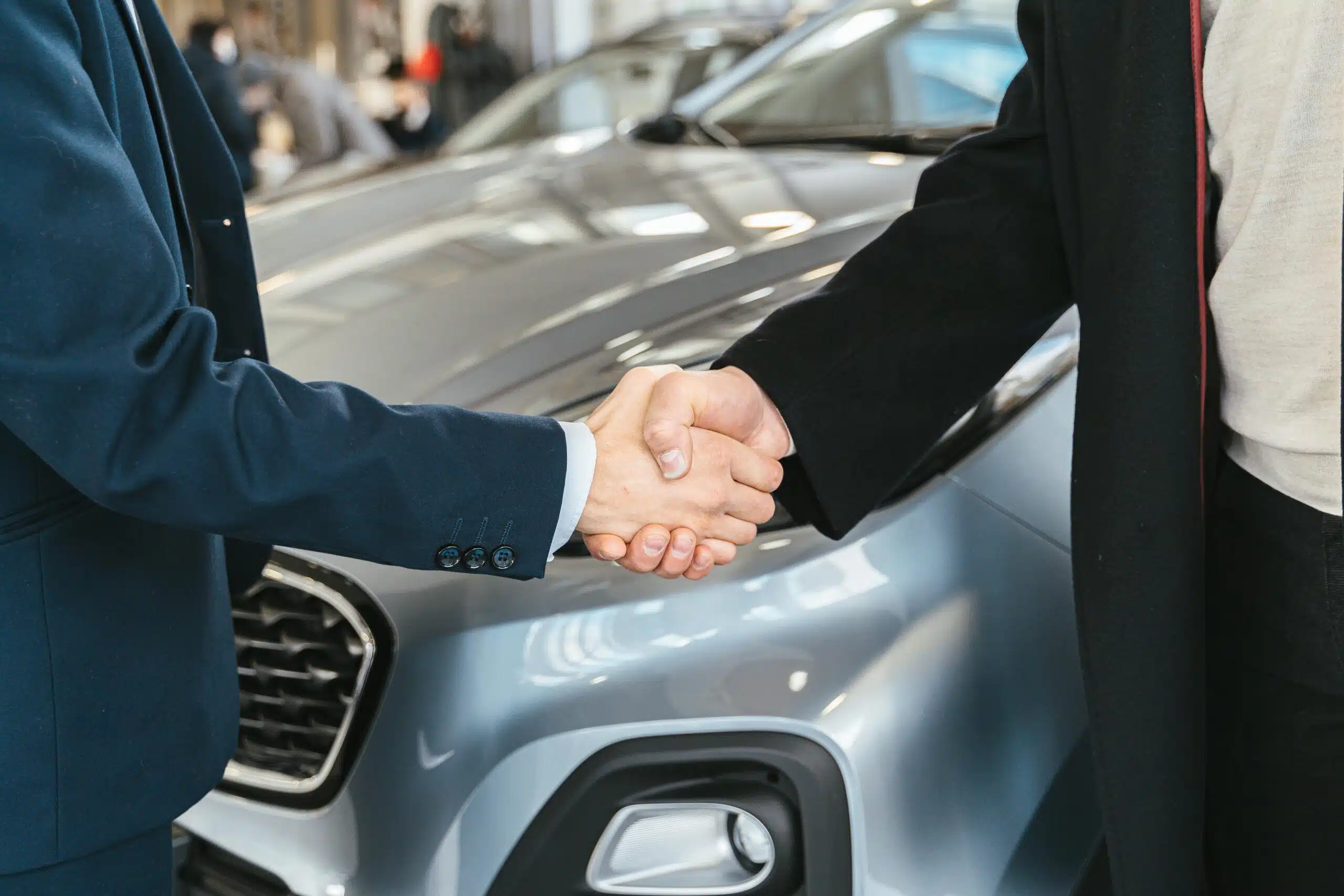 Two people in formal attire shake hands in front of a grey car, marking the culmination of buying a new car at the dealership during Christmas.