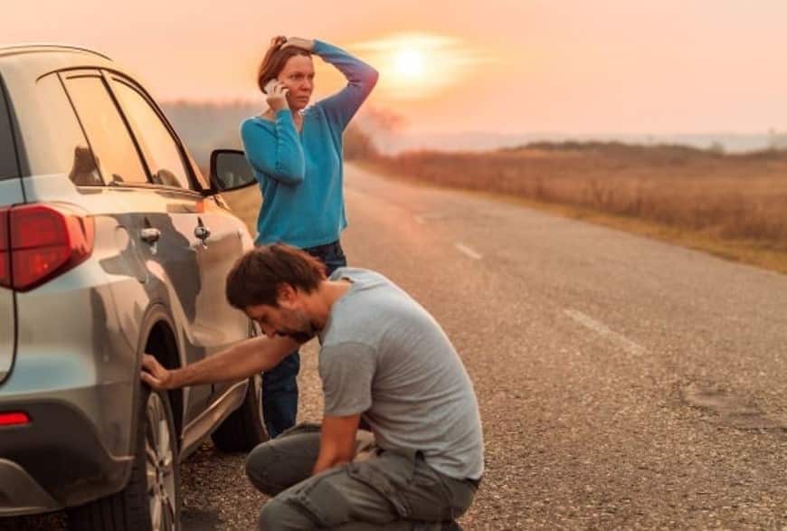 couple on side of road with tyre puncture at sunset, woman on phone and man fixing tyre