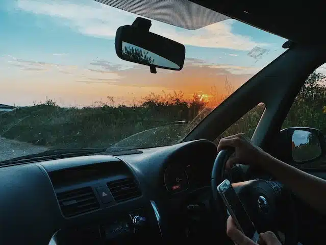 Man behind the steering wheel of a broken down car checks his phone battery while waiting for roadside assistance