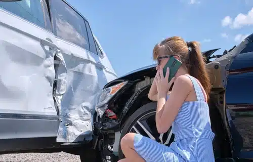 A woman stands beside her car with a damaged door after a car crash, talking on the phone to figure out what to do after a car accident.