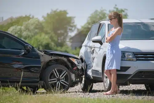 A woman stands with crossed arms next to two damaged vehicles.