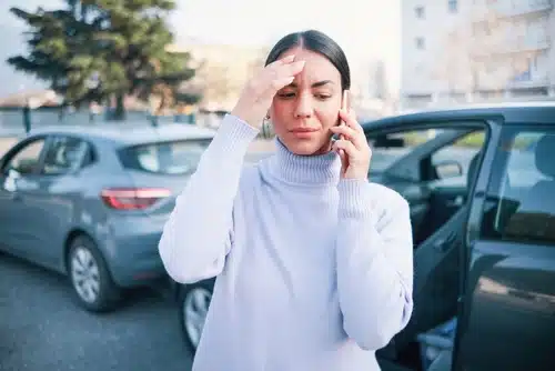 An insured woman standing next to her car, discussing a claim on the phone.
