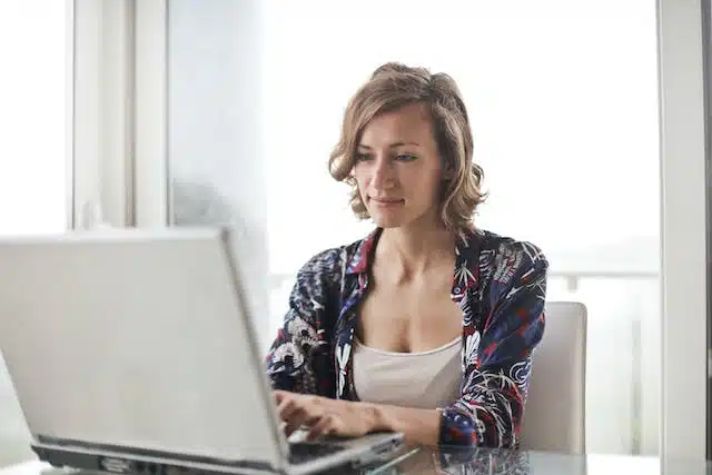 A woman sitting at a table using a laptop to process her car insurance claim after being involved in an at-fault car accident where the other driver doesn't have insurance.