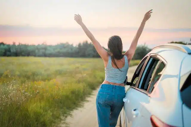 A woman takes a break in the countryside during a road trip and stretches her arms while thinking "I love my summer car!"