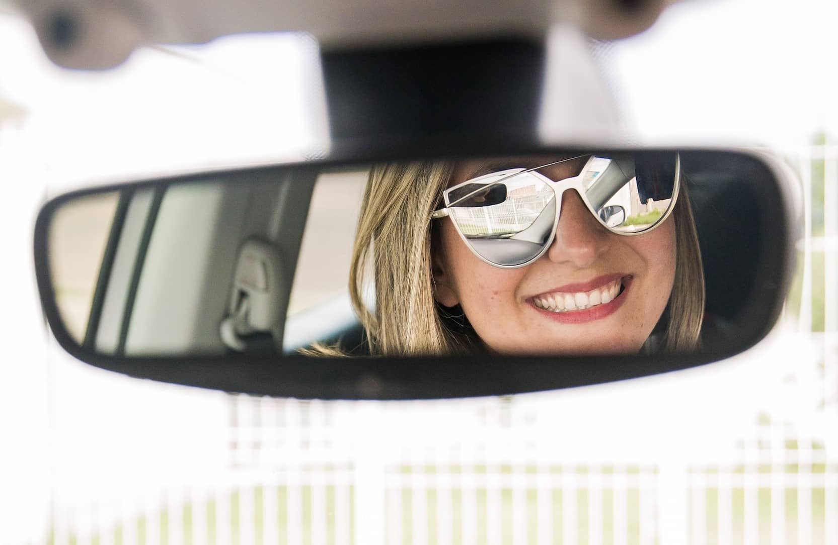 A woman wearing sunglasses in the rear view mirror of a car, plays a car themed game on her phone while waiting for her friend to get ready for a girls' beach trip