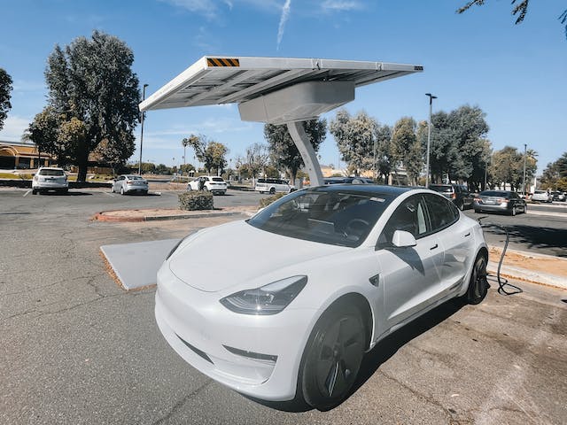 A white tesla model 3 parked next to a solar panel charging station which was bought and installed in part with a government rebate for electric cars' recharging stations
