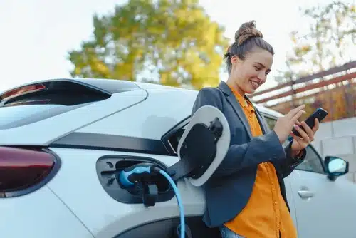 A woman is using her phone while charging her electric car, happy to have taking advantage of the government rebate.