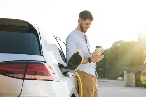 A man is charging his smart car while doing work on his phone over a cup of takeaway coffee