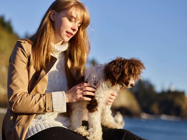 A woman sits on a rock by the ocean with her labradoodle puppy.