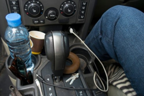 A person sitting in the driver's seat of a messy car with cables, water bottle and old takeaway cup