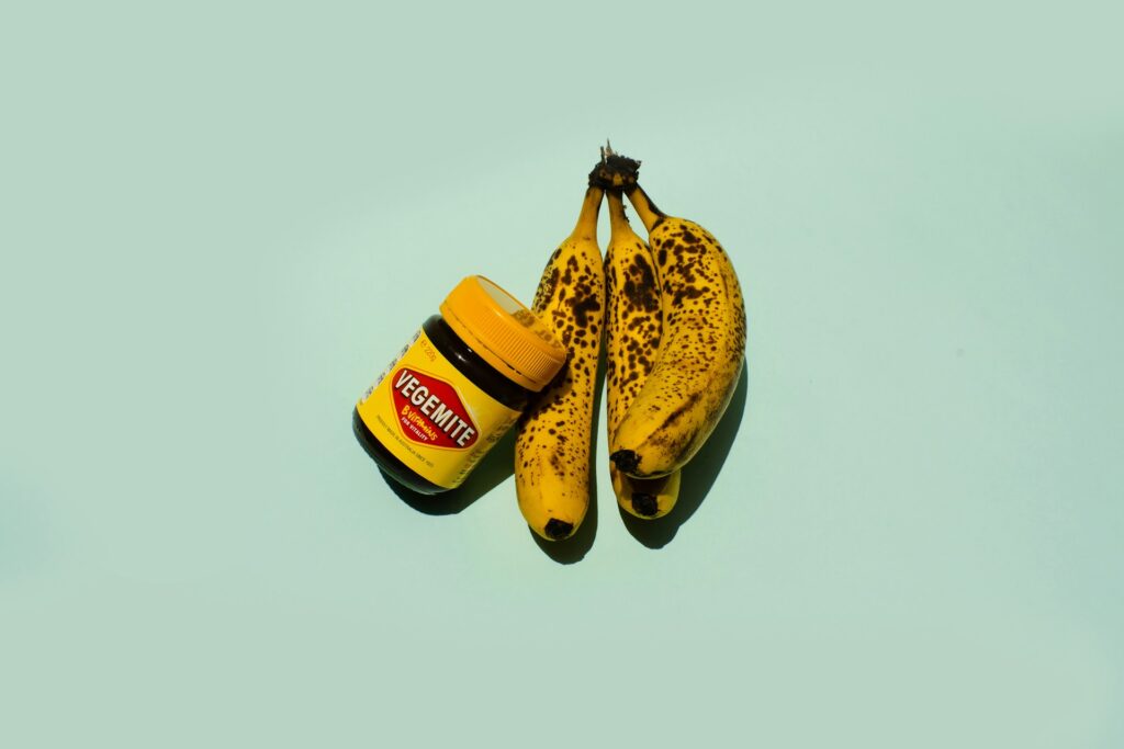Two bananas and a jar of Vegemite on a blue surface.
