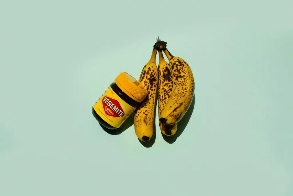 Two bananas and a jar of Vegemite on a blue surface.