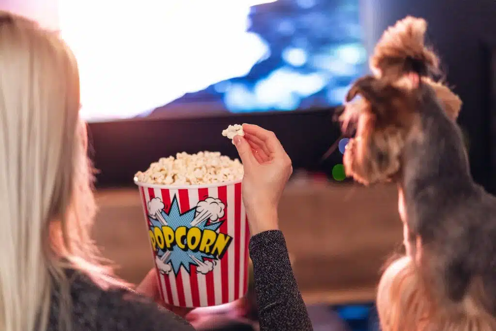 A woman is enjoying popcorn with a dog in front of a TV.