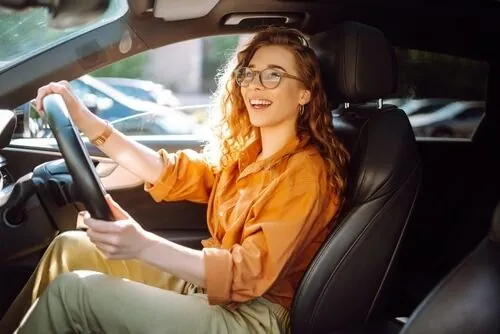 A smiling woman with glasses follows a checklist before starting a car, driving it in daylight.