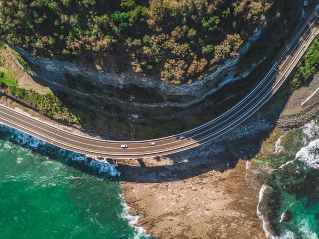 Aerial view of the Great Ocean Road curving along a cliffside with cars driving, green ocean waves below, and dense vegetation above. Perfect for an Easter road trip with breathtaking views that charm any traveler.