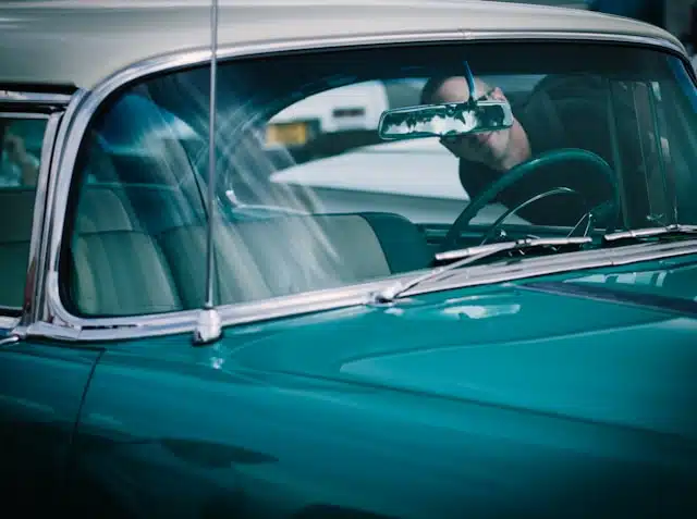 Man driving a vintage teal car seen through the side window with reflection in rearview mirror, pondering car insurance questions.