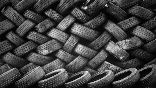 Black and white photograph of a pile of used car tyres densely stacked in various orientations.